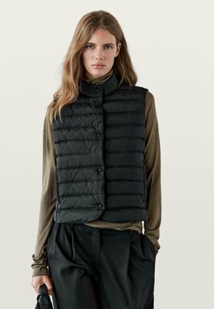 Zwarte donsbodywarmer met doorgestikt ontwerp, hoge kraag en knoopsluiting aan de voorkant, gedragen over een olijfgroen shirt met lange mouwen en losse zwarte broek.