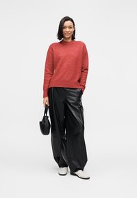 Pull en laine rouge, pantalon large en similicuir noir, et chaussures blanches à enfiler. Accessoires comprenant un petit sac à main noir.