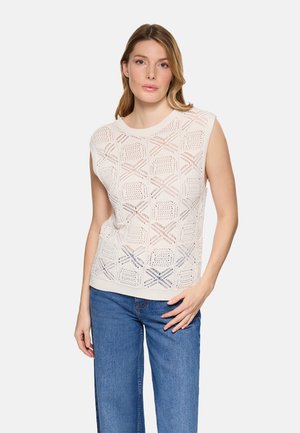 Mujer con top de punto blanco sin mangas con patrones geométricos y jeans azules, posando frente a un fondo claro y liso.