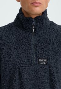 Pullover in pile navy con colletto alto, zip parziale e finitura morbida e testurizzata. Presenta un'etichetta con logo ricamato sul petto.