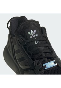 Black athletic shoe featuring mesh and suede materials, textured black laces, and a prominent iridescent metal eyelet detail.