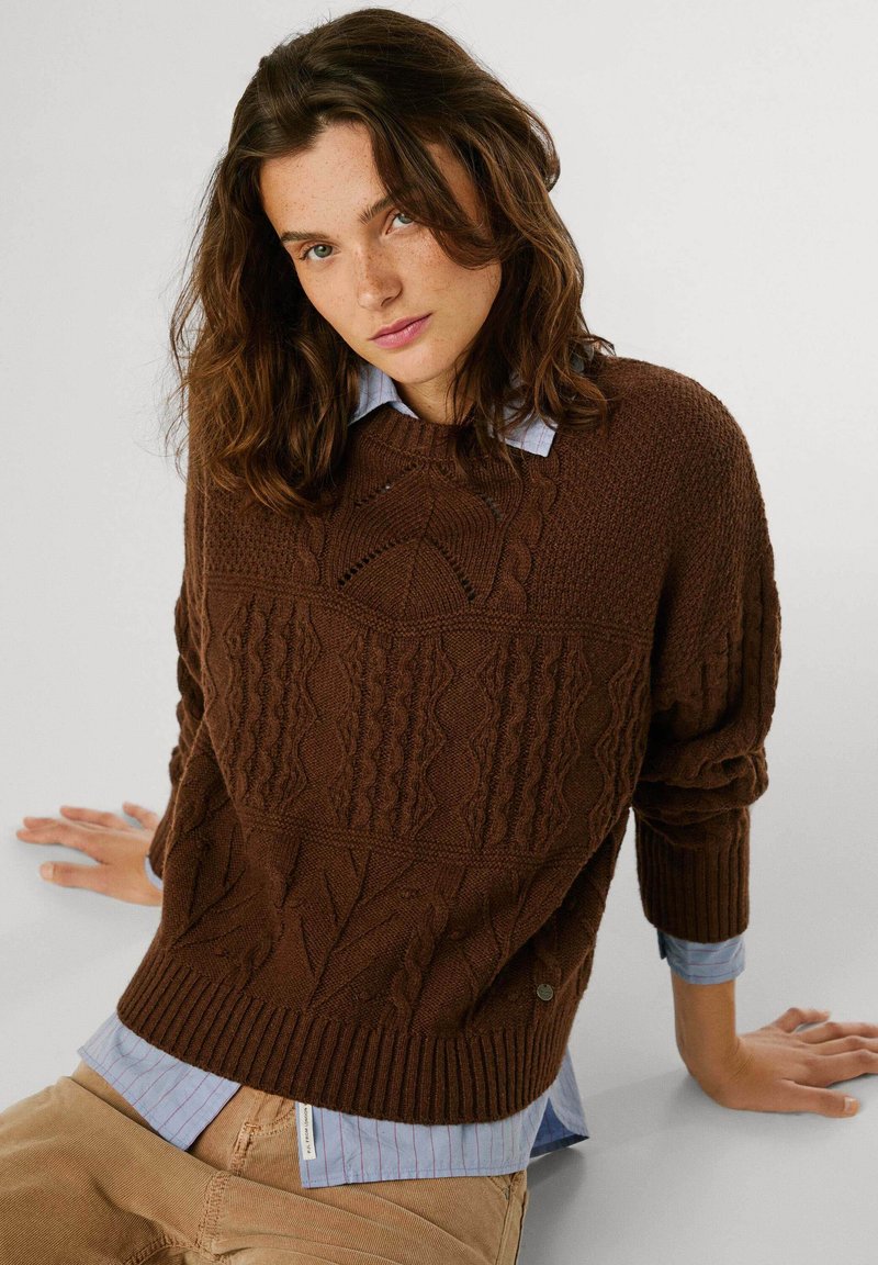 Maglione marrone a maglia con motivi a treccia testurizzati, polsini e orlo a coste. Abbinato a una camicia colletto azzurro chiaro, con righe verticali.