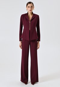 Femme portant un tailleur pantalon violet foncé avec une veste zippée, un pantalon large, des boucles d'oreilles en or, et des chaussures pointues bordeaux, se tenant de face.