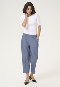 Pantaloni grigi chiari su misura con tasche laterali e una vestibilità rilassata, abbinati a una maglietta bianca aderente. Scarpe con tacco nero e fiocchi.