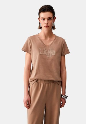 Giovane donna che indossa una maglietta marrone a scollo a V con il logo "Liu Jo" e pantaloni abbinati, accessoriata con grandi orecchini e braccialetti argentati.