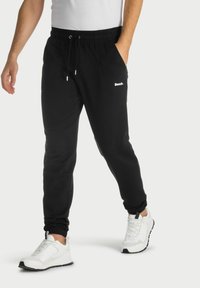 Pantalon de jogging noir avec taille élastique, poches latérales et détails de logo blanc. Associé à des chaussures de sport blanches. Tissu au toucher lisse.
