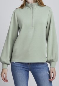 Jersey de color verde claro con cuello alto, cierre frontal de medio cremallera y mangas abullonadas. Hecho de material suave con una textura lisa.