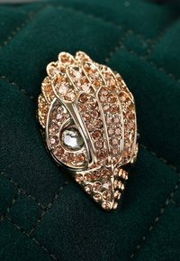 Złoty broszka z misternymi wzorami, wysadzana jasnoróżowymi i przezroczystymi kryształami, spoczywająca na ciemnozielonym pikowanym materiale.