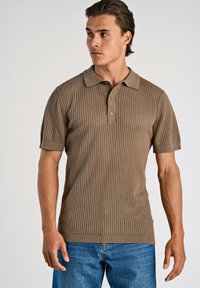 Khaki kortärmad polo med texturerad stickning och krage, med treknappsslutning och ribbad nederkant, bärs tillsammans med blå jeans.