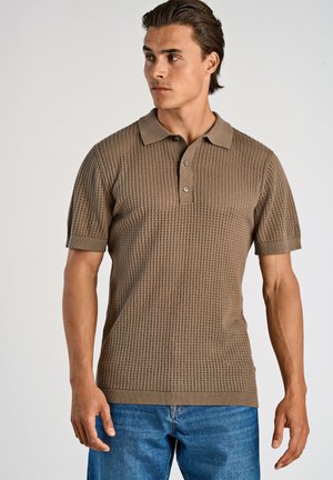 Camisa polo de manga corta color caqui con tejido texturizado y cuello, con tapeta de tres botones y dobladillo acanalado, combinada con unos jeans azules.