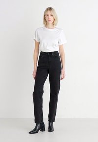 Jean noir taille haute avec une finition texturée, associé à un t-shirt blanc ajusté et des bottines noires, se tenant devant un fond uni.