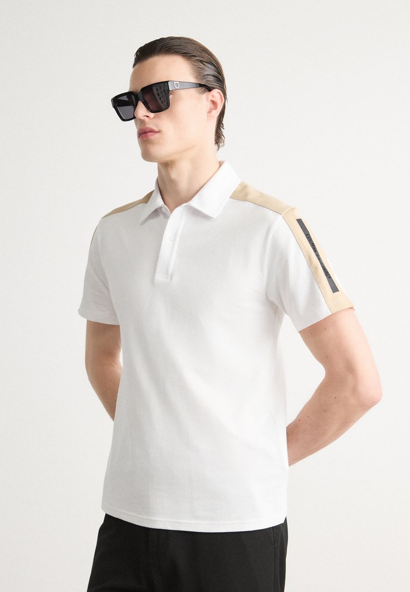 Balta polo marškiniai su smėlio spalvos audiniu ant pečių, trumpomis rankovėmis ir trijų mygtukų užsegimu. Juodi akcentai ant rankovių, lygi medvilnės tekstūra.