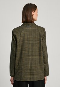 Blazer verde a quadri con maniche lunghe, texture simile alla lana, rever a punta, dettaglio con spacchetto posteriore e una piccola etichetta del marchio sul collo.