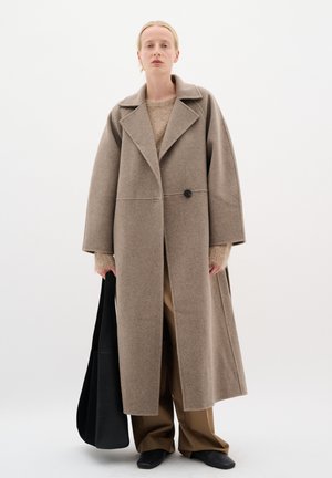 Cappotto lungo e oversize in lana beige con collo ampio e chiusura a singolo bottone. Abbinato a un maglione e pantaloni beige, con una borsa a tracolla nera.