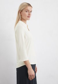 Cream-colored, button-up shirt with a collar, three-quarter sleeves, soft texture, and a relaxed fit. Worn with a dark skirt.