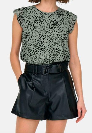 Vrouw draagt een groene top zonder mouwen met stippen en gerimpelde schouders, weggestopt in hoogwaist zwarte leren korte broek met een brede riem.