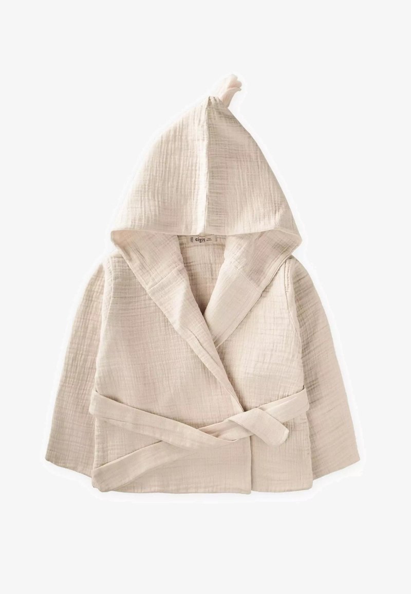 Svetlo béžový plášť s kapucňou vyrobený z mäkkej, textúrovanej tkaniny; má prekrývajúci sa dizajn, viazací opasok a roztomilý uzlík na kapucni.
