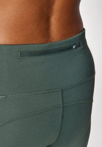 Collant atletici verde scuro con una texture liscia, dotati di una tasca con zip e cuciture visibili lungo le cuciture.
