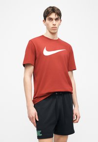 Νέος άνδρας που φοράει κόκκινο T-shirt Nike και μαύρο σορτς με κορδόνι, στέκεται μπροστά σε απλό λευκό φόντο.