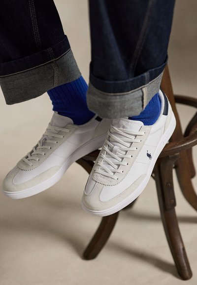 Vita sneakers med snören och marinblå polo-logga, använda med knallblå strumpor och uppkavlade mörka jeans, vilande på en trästol.