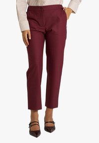 Pantaloni burgundy su misura con una consistenza liscia e una gamba dritta, caratterizzati da una chiusura frontale con bottoni e tasche laterali. Indossati con scarpe nere a punta.