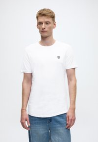 Bijela pamučna majica s okruglim izrezom i kratkim rukavima. Na lijevoj strani prsa nalazi se mali crni logotip u obliku štita. Kombinirana s traper kratkim hlačama.