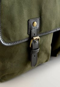 Oliivinvihreä kanvaslaukku tummalla nahkayksityiskohdalla, jossa on messinkinen solki, tikatut reunat ja jämäkkä läppädesign.