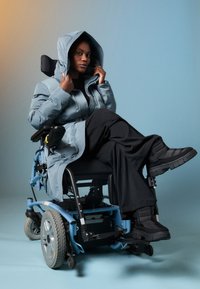 Cămașă umplută de culoare albastru deschis cu glugă, pantaloni negri texturați și pantofi chunkie negri, așezat într-un scaun cu rotile electric albastru, pe un fundal gradient.