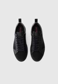 Pair of black low-top sneakers with black laces, gray textile collar, and red "HUGO" logo on insoles and sides, displayed on white background.