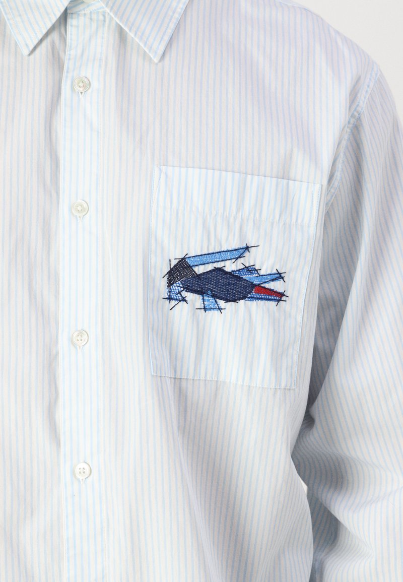 Camicia a righe blu chiaro con taschino sul petto, caratterizzata da un design ricamato in blu e rosso, che presenta forme geometriche e texture.