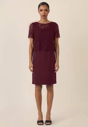 Frau steht, trägt ein knielanges bordeauxrotes Kleid mit Spitzenüberzug oben und kurzen Ärmeln, kombiniert mit schwarzen, offenen High-Heels vor einem schlichten Hintergrund.