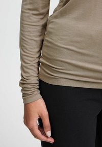 Top de manga larga ajustado en suave tejido beige, con una textura lisa y detalle fruncido en la cintura, combinado con pantalones negros.