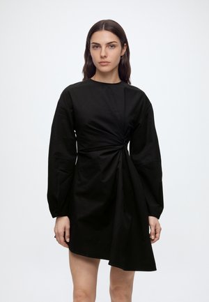 Femme aux longs cheveux foncés portant une robe noire asymétrique à manches longues avec un détail froncé sur un côté, debout devant un fond uni.