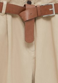 Pantalons beiges avec une finition texturée et une ceinture en cuir marron. La ceinture présente une boucle en métal et est passée à travers les passants de ceinture.