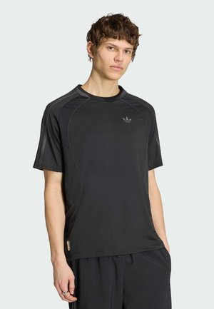 Ung mand iført en sort Adidas sportstrøje med korte ærmer og sorte bukser, stående mod en ensfarvet lys baggrund.