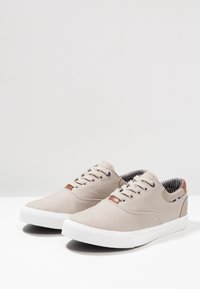Beiga canvas-sneakers med vita gummisulor, rund tå och bruna läderdetaljer; fodrade med randig tyg vid öppningen.
