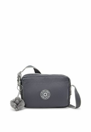 Sac bandoulière gris avec poche zippée à l'avant, patch rond avec logo Kipling et un petit porte-clés singe en peluche attaché à la sangle.