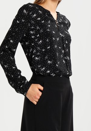 Femme portant un chemisier noir avec un motif d'insectes blancs et de points, rentrée dans un pantalon noir taille haute, main dans la poche.