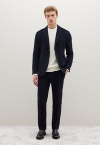Completo in velluto a coste blu navy con blazer strutturato e pantaloni abbinati. Il modello indossa un maglione color crema sotto. Scarpe nere completano il look.