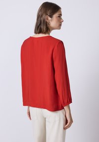 Blusa rossa leggera con maniche lunghe leggermente a sbuffo e scollo rotondo. Il tessuto ha una consistenza liscia e il retro è semplice.