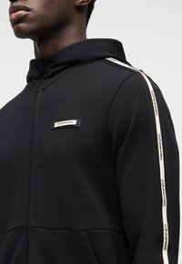 Μαύρο φούτερ με φερμουάρ, κατασκευασμένο από ομαλό ύφασμα, με κουκούλα, πλαϊνές τσέπες και λογότυπο στο στήθος, διακοσμημένο με αντίθετες λεπτομέρειες ταινίας στο πλάι.