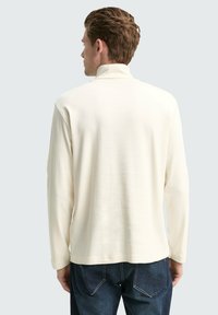 Mann mit kurzem braunem Haar, der einen cremefarbenen Langarmrollkragenpullover und dunkelblaue Jeans trägt und mit dem Rücken zu einer einfarbigen Hintergrund steht.