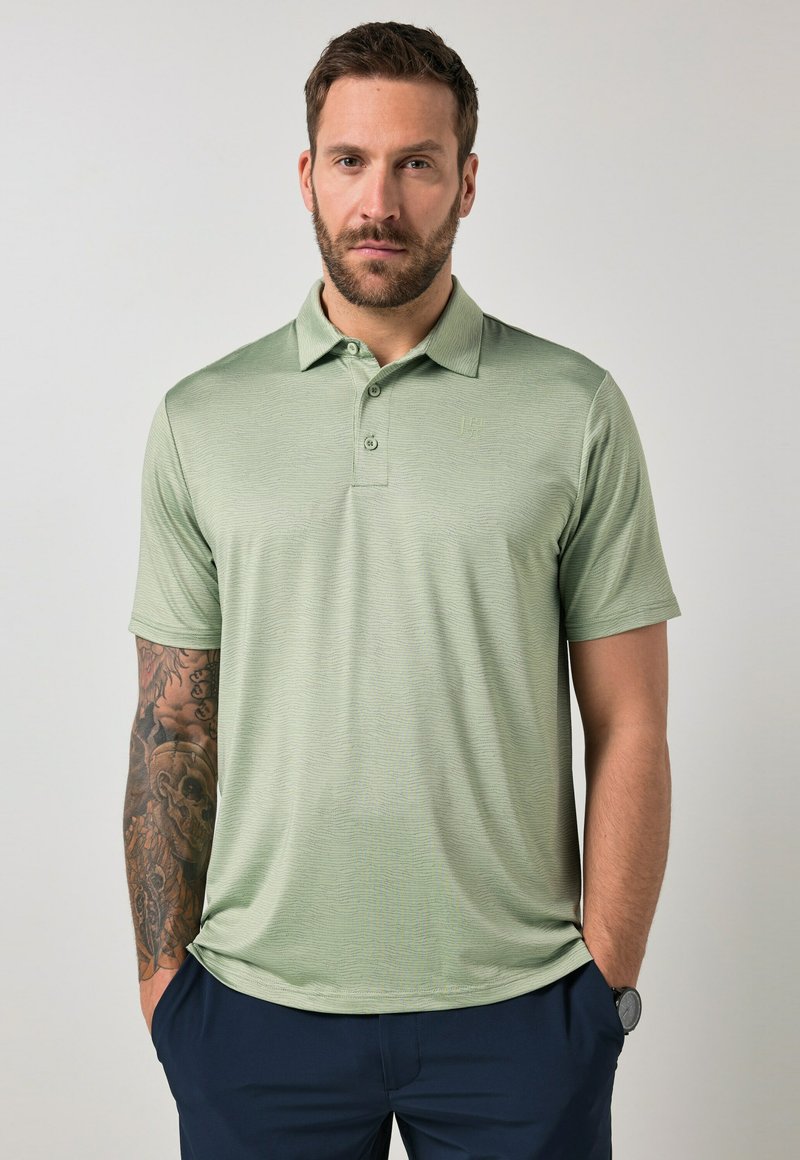 Hombre con barba vistiendo una camisa polo de manga corta verde claro y pantalones azul marino, con un tatuaje que cubre todo el brazo izquierdo y las manos en los bolsillos.