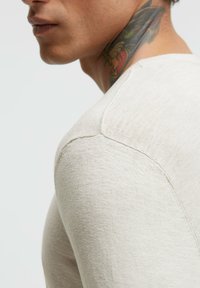 Hellbeige, strukturierter Strickshirt mit rundem Ausschnitt, sichtbaren Nahtdetails und einem exponierten Schulternaht, präsentiert an einem Model.