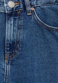 Gros plan sur un jean en denim bleu montrant la poche avant, la passante de ceinture, le bouton en métal, le rivet et les détails des coutures orange.