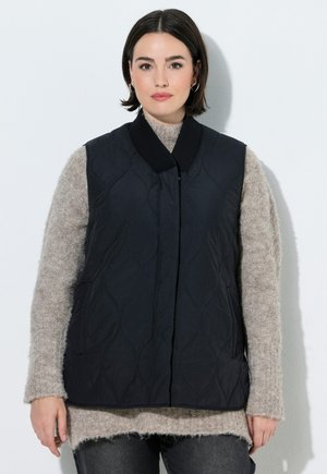 Donna con capelli corti e scuri che indossa un gilet nero trapuntato sopra un maglione beige peloso e pantaloni scuri, in piedi davanti a uno sfondo semplice.