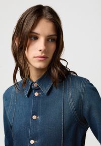 Camisa de denim con cuello alto, cierre de botones y costuras visibles. Color azul oscuro con una textura suave y cuatro botones prominentes.