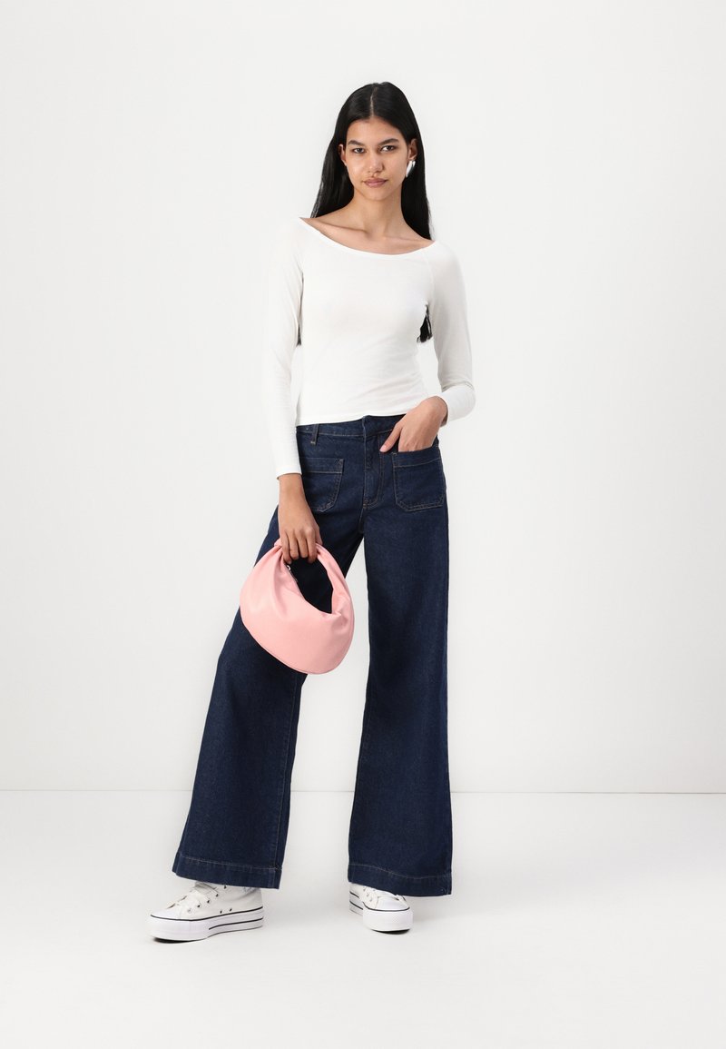 Top a maniche lunghe bianco, jeans a gamba larga blu scuro e sneakers bianche. Tieni in mano una borsa rosa, presentata su uno sfondo chiaro e uniforme.