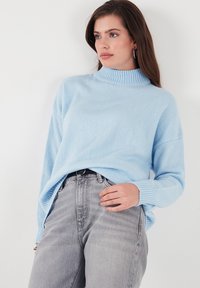 Gaiši zils pārāk liels džemperis ar augstu ribotu apkakli un vaļīgām piedurknēm, saskaņots ar pelēkiem augstām jostasvietas džinsiem. Mīksta adījuma tekstūra.