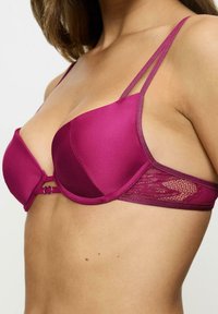 Reggiseno magenta in satin con ferretto, dettagli in pizzo sui fianchi e spalline sottili regolabili. Presenta una chiusura a gancio davanti.